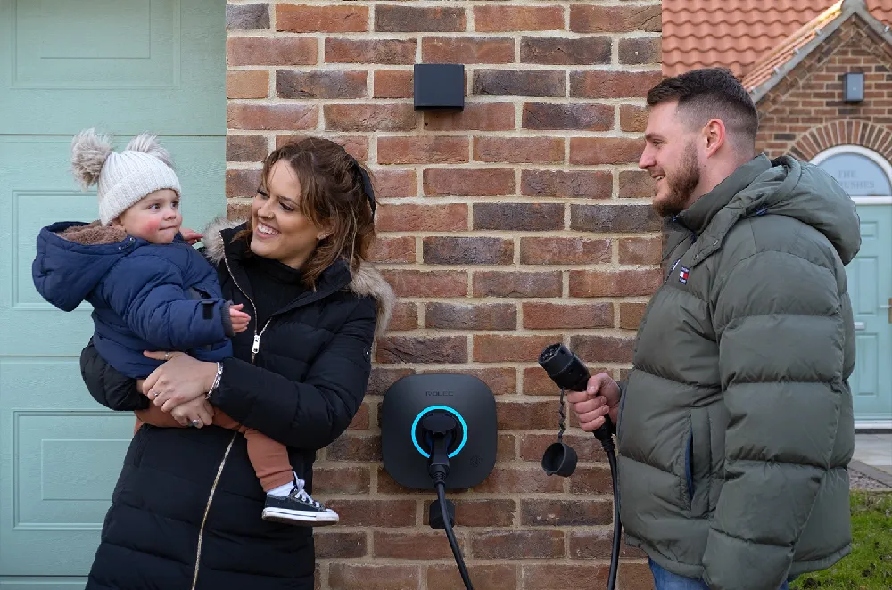 A family stands outside their home next to a wall-mounted Rolec EVO EV charger. A woman holds a smiling toddler in winter clothing, while a man in a green puffer jacket holds the charger plug. The charger is installed on a red brick wall, glowing blue, with a mint green garage door and house entrance in the background.